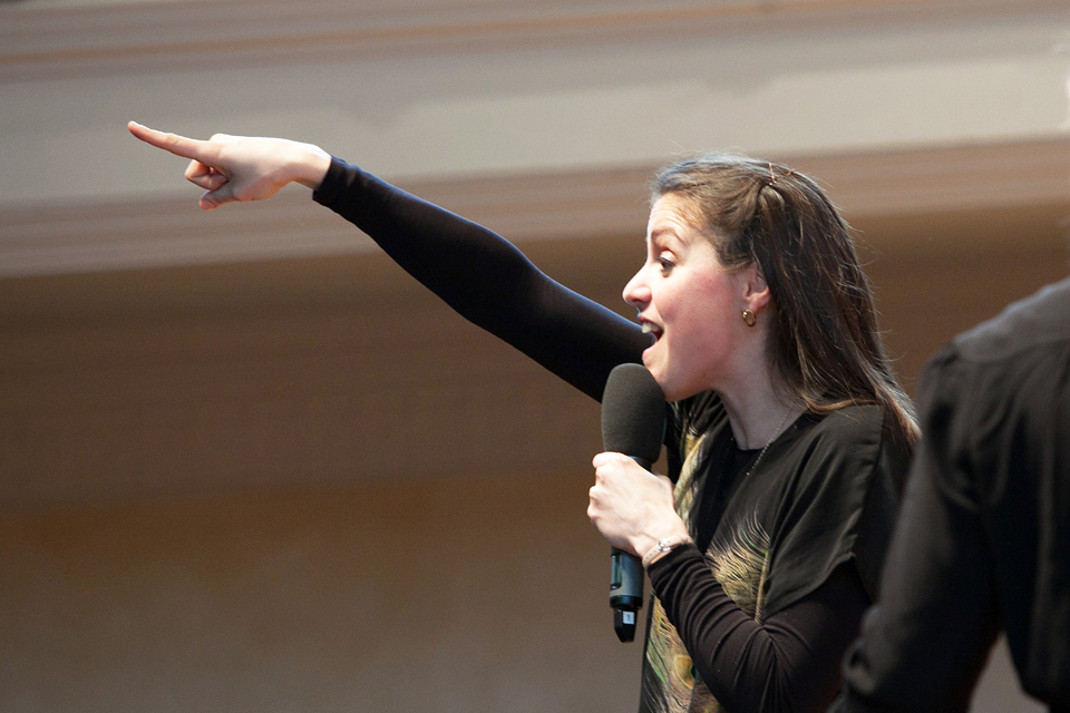 A women, with dark hair, speaking into a microphone and pointing out into the audience.